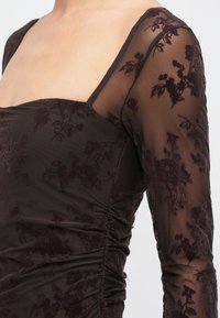 Dunkelbraunes Kleid mit quadratischem Ausschnitt, durchsichtigen Mesh-Ärmeln, floralen Samtmustern und gerafften Stoffdetails an der Taille.