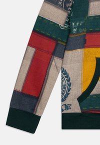Pull en patchwork avec des sections rouges, vertes, navy et jaunes. Poignets et ourlet côtelés en vert foncé ; tissu texturé.
