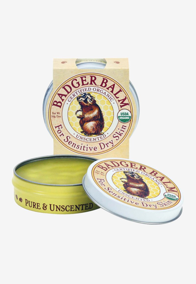 Badger Balm metallisen pyöreän tinan sisällä, jossa on keltainen etiketti, jossa on karhun grafiikka, ja teksti "Hajusteeton" sekä "Herkkäihoiselle kuivalle iholle".