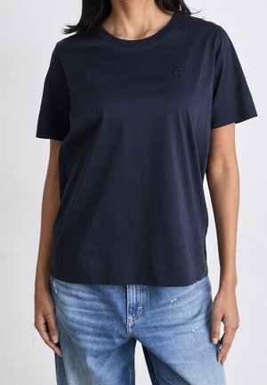 Osoba ubrana w gładki, ciemnogranatowy t-shirt z krótkim rękawem i jasnoniebieskie dżinsy, stojąca z ramionami swobodnie opuszczonymi wzdłuż ciała.