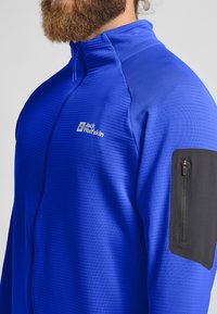 Blaue Sportjacke aus strukturiertem Stoff, mit Viertel-Reißverschluss-Design, kontrastierendem schwarzen Reißverschlussfach am linken Ärmel und Logo auf der Brust.