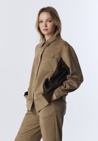 Vrouw met blond haar draagt een tan suède jack en bijpassende broek, poseert met handen in de zakken van het jack tegen een effen lichte achtergrond.