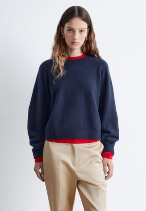 Navy oversized sweater with a textured finish and ribbed cuffs, layered over a red top, paired with light beige trousers.