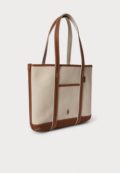 Μπεζ τσάντα ώμου από καμβά με καφέ δερμάτινες λεπτομέρειες, ραφές σε αντίθεση και μπροστινή τσέπη. Διαθέτει ανθεκτικούς ιμάντες ώμου και λεπτομέρεια με λογότυπο.