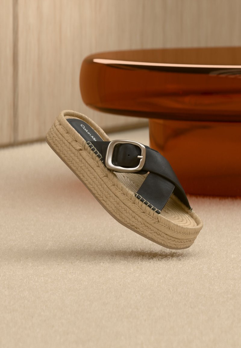 Sandale à plateforme avec semelle en jute tressée, lanières croisées en cuir noir et grande boucle argentée, placée sur un tapis beige et une table en bois.