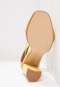 Żółte zamszowe sandały na wysokim obcasie z szeroką podstawą, otwartym noskiem i regulowanym paskiem wokół kostki; gładkie wewnętrzne wykończenie z teksturowaną podeszwą.