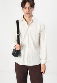 Gestreiftes, cremefarbenes Langarmhemd mit einem Button-Down-Kragen; kombiniert mit braunen Hosen; hält eine kleine schwarze Umhängetasche.