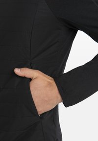 Black jacket featuring a textured front with horizontal lines, side pocket with zipper, and snug black sleeves. Soft fabric with a matte finish.