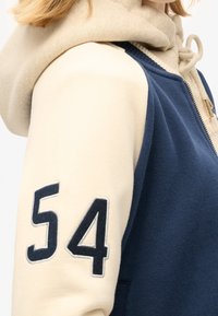 Granatowo-kremowa bluza rozpinana na zamek z haftowanym numerem "54" na lewym rękawie, o miękkiej fakturze i z kapturem.