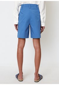 Blå bomuldsshorts med lige snit, forlommer og en enkelt baglomme. Kombineret med flettede sandaler. Billedet viser bagsiden.