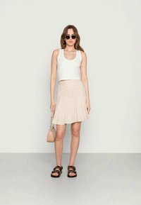 Top cropped branco, saia rodada bege com padrão xadrez, sandálias pretas e uma pequena bolsa bege. Modelo em pé sobre um chão cinza claro.