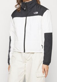 Kurtka puchowa z białym korpusem i czarnymi ramionami oraz rękawami. Zamek błyskawiczny z przodu, wysoki kołnierz i poziome przeszycia dla uzyskania tekstury.