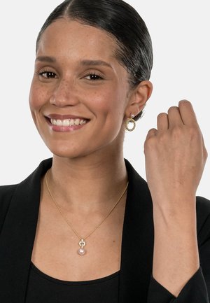 Goldkette mit einer Perlenanhänger, getragen mit passenden goldenen Creolen, an einer Person in einem schwarzen Oberteil und Blazer. Die Haut ist hell mit Sommersprossen.