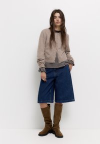 Beige kötött kardigán gombos elől, egy fekete-fehér csíkos ing fölött. Kék farmer culotte és barna térd fölé érő csizma egészíti ki az outfitt.