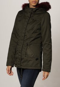 Kobieta w oliwkowej parze kurtki z burgundowym futerkiem na kapture, z kieszeniami z przodu i regulowanym pasem, zestawionej z ciemnoniebieskimi dżinsami.