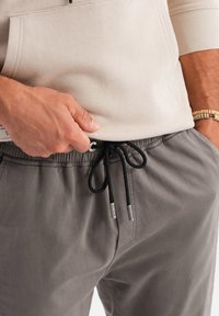 Graue verstellbare Hosen mit einem Kordelzugbund. Der Kordelzug hat Metallenden. Der Stoff hat eine glatte Textur und einen lockeren Schnitt.
