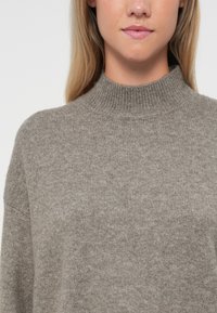 Taupe høyhalset genser laget av mykt strikket stoff. Har høy krage og avslappet passform, med en jevn tekstur og subtil variasjon i fargen.