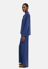 Ciemnoniebieska, długosleevowa bluzka z klapami oraz dopasowane szerokie spodnie wykonane z miękkiej, teksturowanej tkaniny, noszone z brązowymi butami.