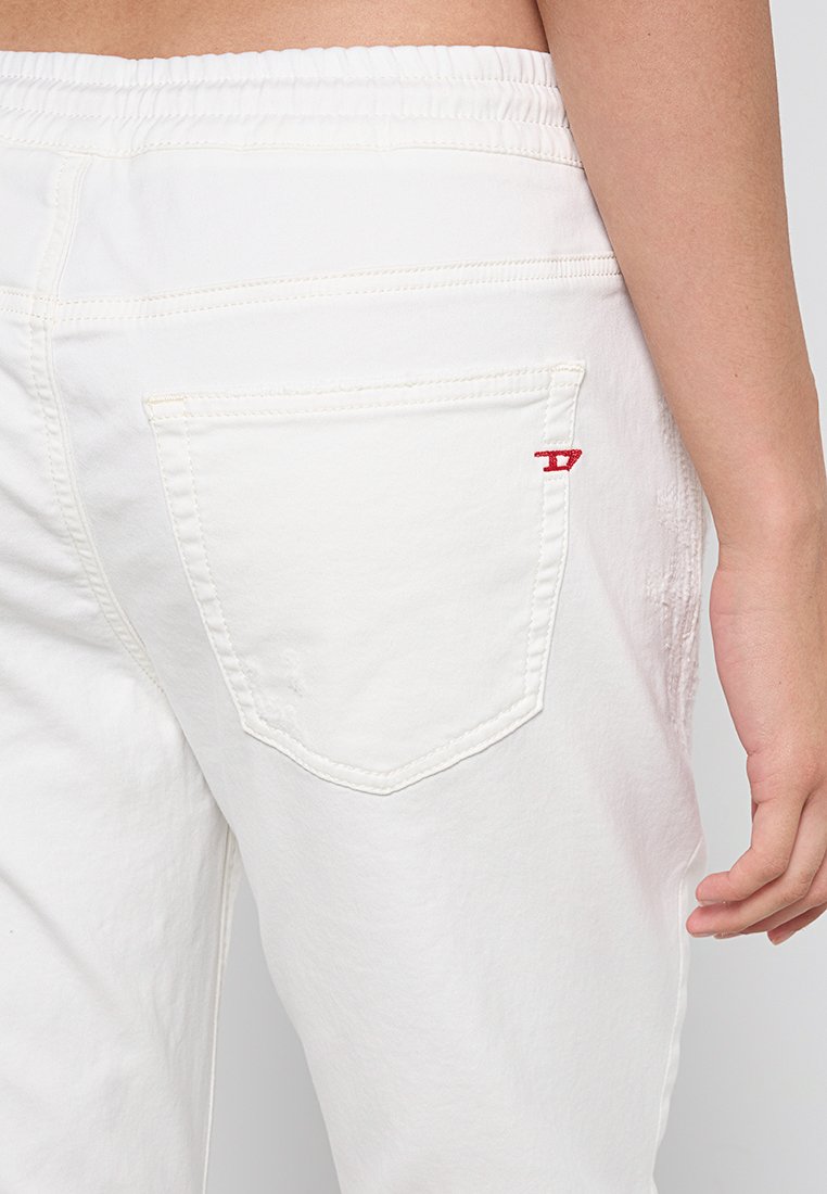 Person, der weiße Hosen mit einem elastischen Bund und einer Gesäßtasche trägt, die ein kleines rotes, gesticktes Detail aufweist.