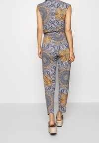 Femme portant une combinaison sans manches à motifs avec des dessins mandala orange, noir et gris, associée à des sandales compensées beiges, debout, de dos.