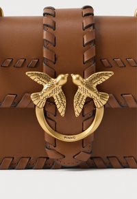 Braune Lederhandtasche mit geflochtenen Akzenten und goldfarbenem rundem Beschlag, der zwei Vogelgestalten zeigt. Kompakte rechteckige Form.