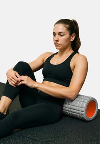Grauer Schaumstoffroller mit strukturierten Oberflächen, orangefarbenes Zentrum, auf einer schwarzen Trainingsmatte positioniert. Die Person sitzt und trägt schwarze Sportbekleidung.
