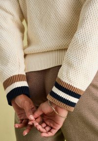 Cremefarbene Strickjacke mit gerippten Bündchen in marineblau, beige und braunen Streifen. Die Hände sind lässig hinter dem Rücken verschränkt, was ein Kettenarmband zeigt.