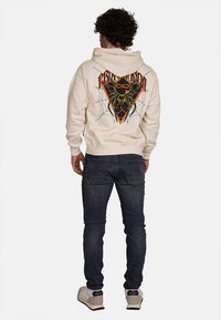 Felpa crema con grafica di ragno e design a ragnatela sul retro, abbinata a jeans blu scuro e sneaker beige, indossata da una figura maschile.