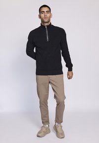 Suéter negro de punto con cremallera y cuello alto, combinado con pantalones beige y zapatillas de color claro, de pie contra un fondo liso.