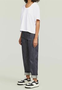 Camiseta blanca corta, de corte holgado, con cuello redondo, combinada con jeans de corte recto en lavado oscuro, con los puños doblados, y zapatillas negras y blancas.