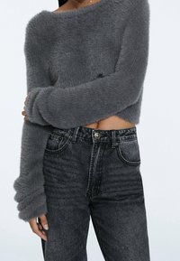 Persona vistiendo un suéter gris afelpado y corto, y unos jeans negros de cintura alta deslavados, con un brazo cruzado sobre el torso.