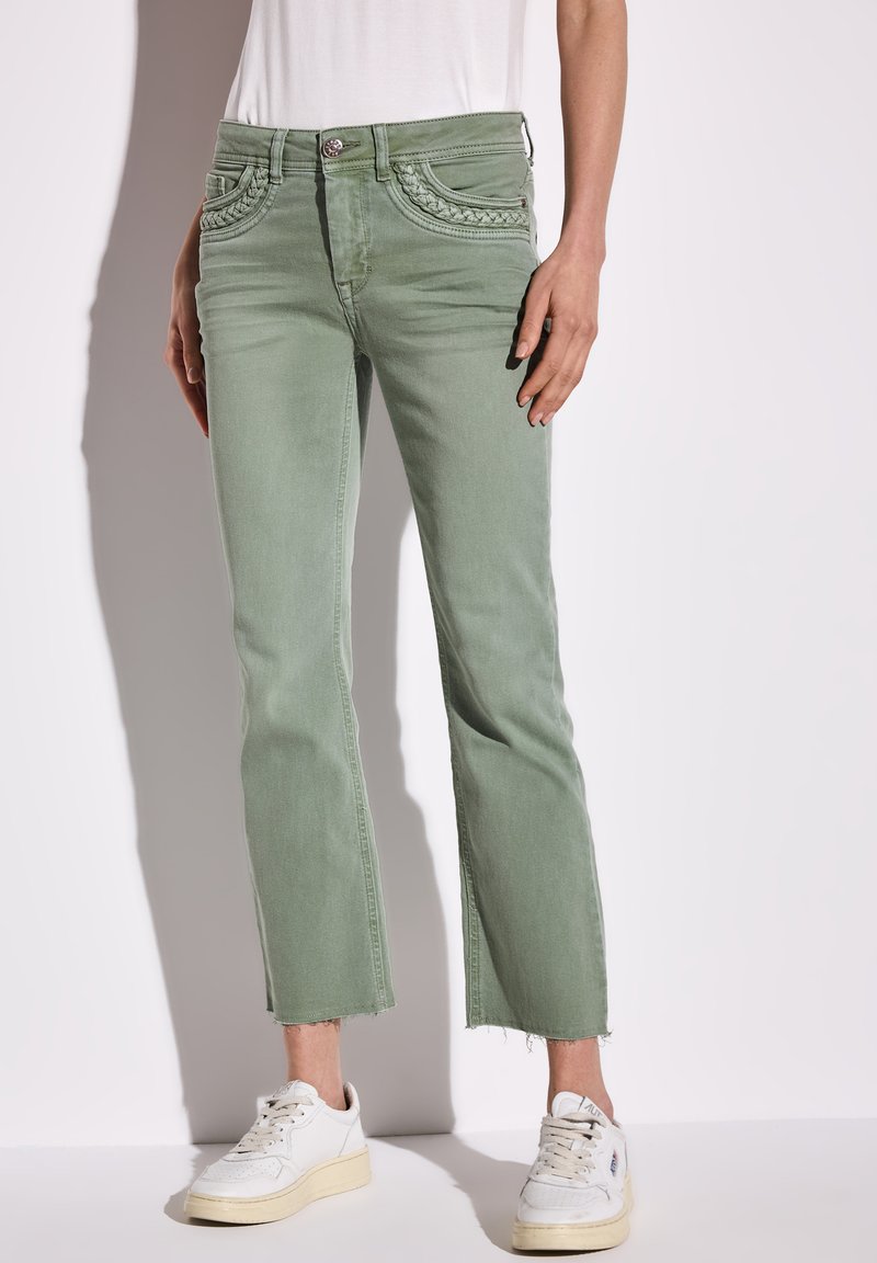 Hellgrüne, verkürzte Jeans mit geflochtenem Taschendetail, ausgefranster Saum und klassischem Fünf-Taschen-Design, kombiniert mit weißen Sneakers.