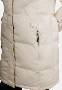 Beige boblejakke med quiltet tekstur, som har en sidelomme med glidelås og en klaff med trykknapp for ekstra detaljer.