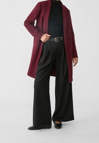 Bordeauxfarbener Wollmantel mit Reverskragen, getragen über einem schwarzen gerippten Rollkragenpullover. Schwarze weite Hosen und schwarze Ballerinas runden das Outfit ab.