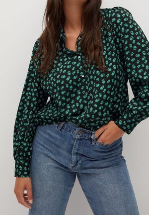 Mujer con blusa negra de botones con patrón floral verde y jeans azules, con una mano en el bolsillo sobre un fondo liso.