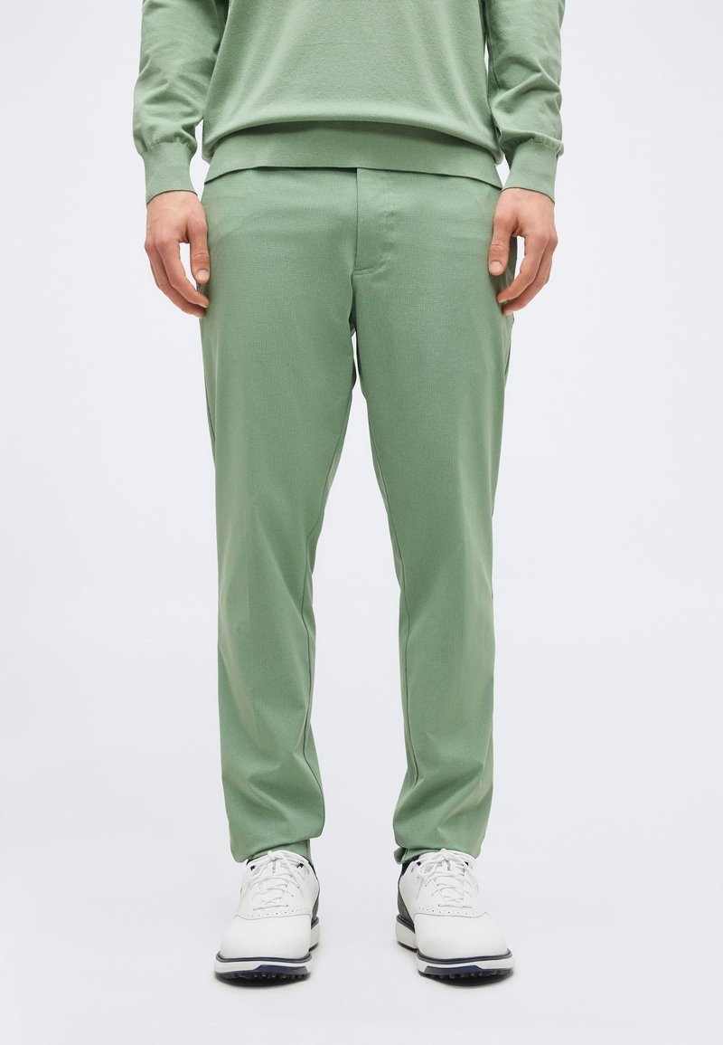 Άνδρας που φοράει ανοιχτοπράσινο παντελόνι, γκριζοπράσινο φούτερ και λευκά δερμάτινα παπούτσια με κορδόνια, στέκεται μπροστά σε απλό φωτεινό φόντο.