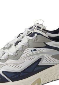 Skriešanas apavi pelēkā, baltā un jūras zilā krāsā. Iespējas ietver teksturētu virsu, acs paneļus un mīkstu zoli ar viļņveida rakstu. Zīmols uz sāniem.