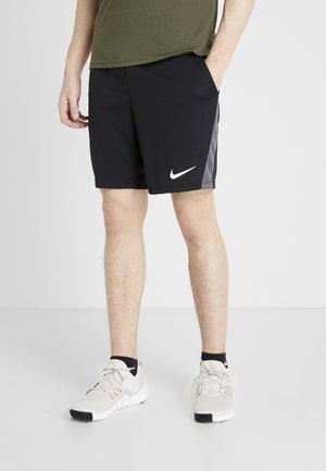 Man draagt zwarte Nike sportshorts met grijze zijpanelen, een olijfgroen shirt, zwarte sokken en witte sportschoenen, staand met één hand in de zak.