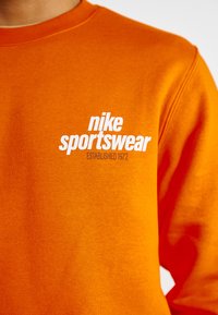 Oranžs Nike sporta krekls ar apaļu kakla izgriezumu un priekšējo baltu logos, pievienots teksts "Izveidots 1972." Redzama teksturēta auduma struktūra.