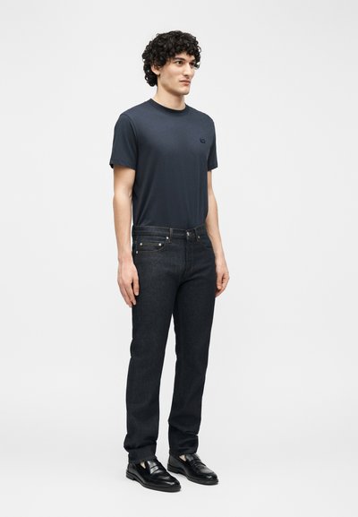 Homme aux cheveux bouclés portant un t-shirt bleu marine foncé, un jean foncé et des chaussures en cuir noires, debout devant un fond clair uni.