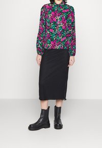 Bloes met lange mouwen in levendig roze en groen op een zwarte achtergrond, gecombineerd met een zwart rok tot de knie en zwarte enkelboots.