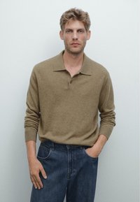 Brauner Strick-Polo-Pullover mit lockerer Passform, langen Ärmeln und Kragen; kombiniert mit dunkelblauen Jeans. Einfarbiger Hintergrund.