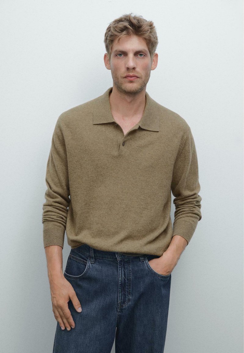 Brauner Strick-Polo-Pullover mit lockerer Passform, langen Ärmeln und Kragen; kombiniert mit dunkelblauen Jeans. Einfarbiger Hintergrund.