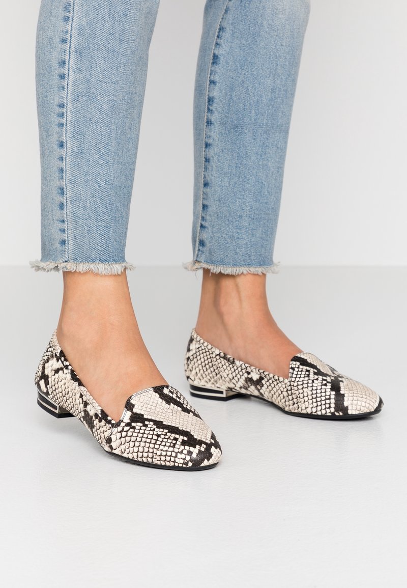 Des pieds portant un jean bleu clair à ourlet effiloché et des mocassins plats à petits talons avec un motif en serpent noir et blanc sur un sol blanc.