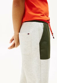 Pantalones de chándal grises con un panel lateral verde, con un bolsillo trasero que lleva un parche con un logo. Cierre de cordón en rojo. Mano de niño visible.