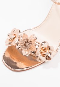 Sandale en or rose métallique avec trois ornements floraux décoratifs sur la bride des orteils et un design à bout carré.