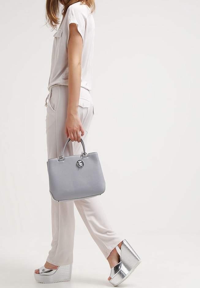 Mujer con un atuendo de color claro caminando, sosteniendo un bolso gris y luciendo sandalias de cuña plateadas sobre un fondo liso.
