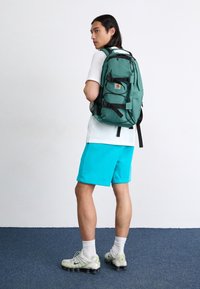Persoon in een wit T-shirt, turquoise shorts, witte sneakers en een groene rugzak staat op een blauw tapijt tegen een witte muur.