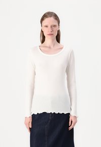 Top bianco a maniche lunghe con scollo tondo e orlo scallop. Texture morbida, design aderente, abbinato a una gonna di denim scura.
