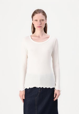 White ribbed long-sleeve top with a round neckline and scalloped hem. Soft texture, fitted design, paired with dark denim skirt.
